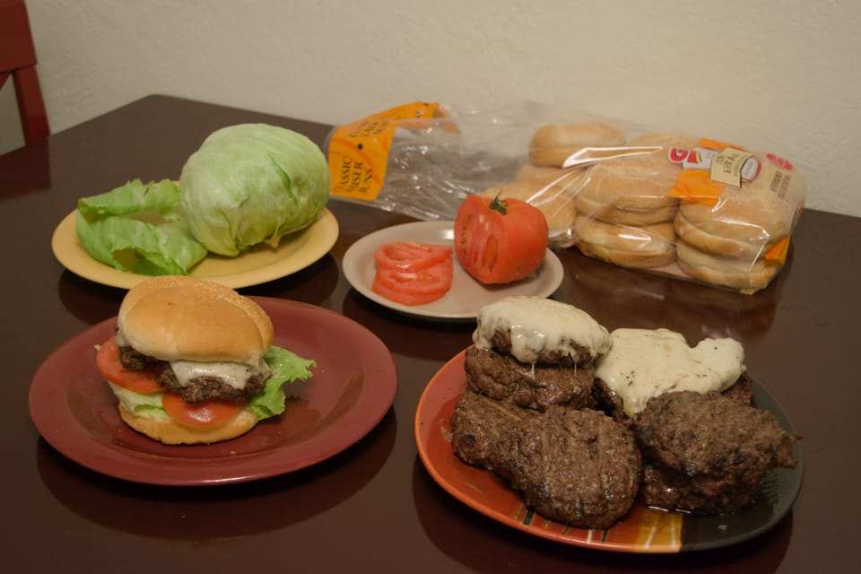 Photo of burgers, buns, tomato, and lettuce on a table.