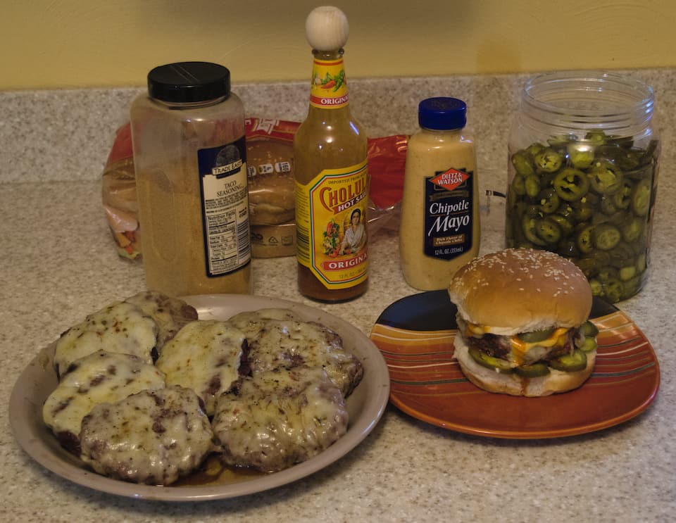 A pile of burger patties, and a finished burger on a bun, with ingredients behind.